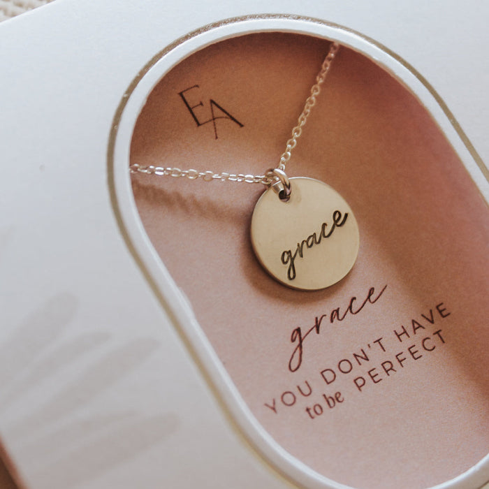 A gold disc pendant necklace engraved with the word 'grace' is displayed inside a white gift box with a peach-colored insert. The insert features the initials 'EA' and the quote 'grace YOU DON'T HAVE to be PERFECT'.